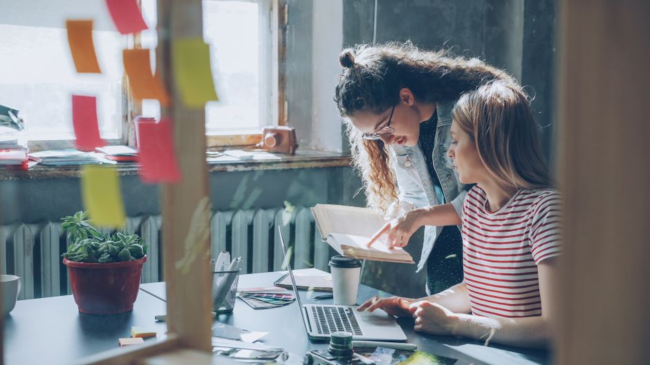 Two women collaborating in a cozy office space with laptops and notes.