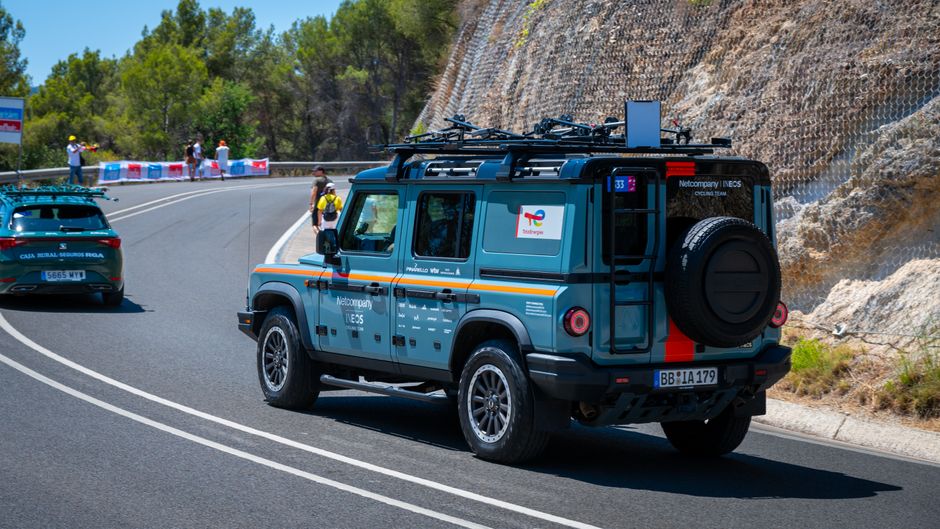 Support vehicles following a cycling event on a winding mountain road under bright sunlight.